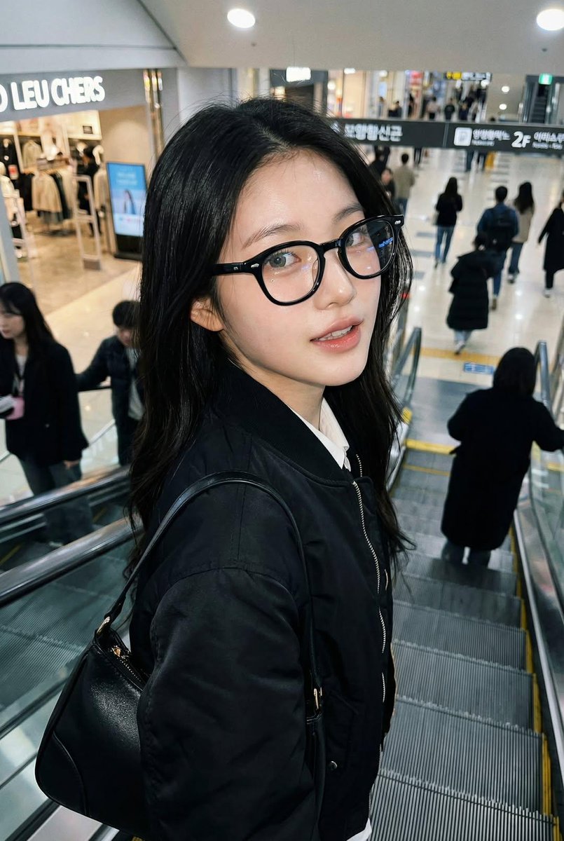 一张像是用 iPhone 拍摄的真实感抓拍照片，画面中是一位女士站在自动扶梯上，面部和身份与参考图完全一致。她戴着黑色玳瑁框眼镜，穿着黑色飞行员夹克，背着一只黑色手提包。姿态自然，处于动作进行...