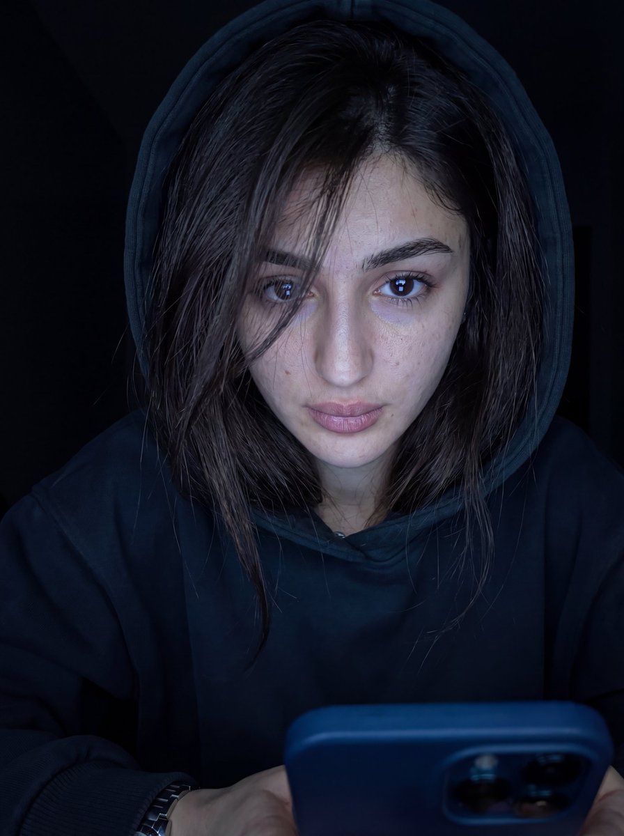 超写实特写肖像，画中人物为男性或女性（来自上传图像），凌乱的头发部分遮住一只眼睛，身穿深色 oversized 帽衫，帽子微微拉起。脸部由下方冷色调的手机屏幕光照亮，眼睛锐利地反射出光线。背景...