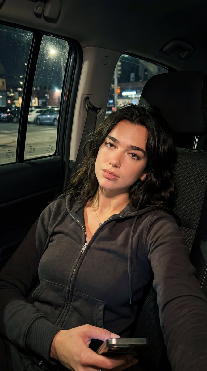 Dua Lipa, 保持面部结构, 手机略低于视线水平,靠在座椅上, 平静,略带魅惑的眼神交流, 深色,松散,略显凌乱, 柔软面料,休闲但有型, 深色修身拉链连帽衫,领口微敞, 车内, 夜晚,...
