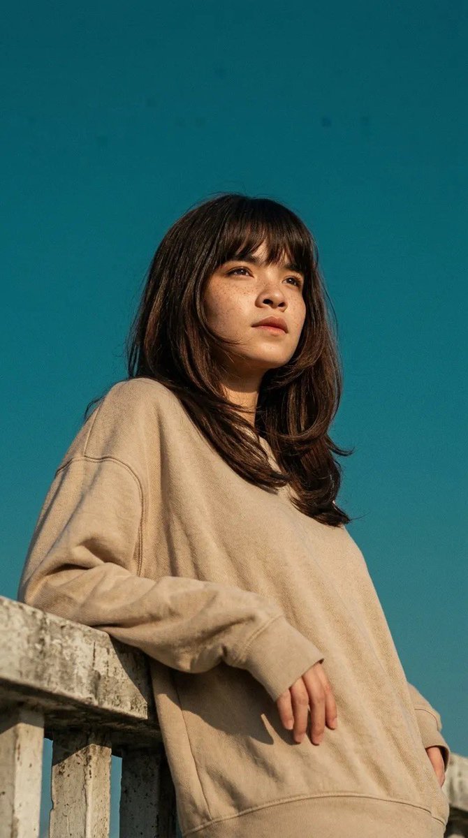 电影感人像照：一位年轻女子倚靠在混凝土栏杆上，身穿宽松米色卫衣，柔和的自然阳光在她脸上投下温暖的色调，背景为清澈的青蓝色天空，淡淡的雀斑，中长深棕色头发配刘海在微风中轻轻飘动，低角度拍摄，极简...