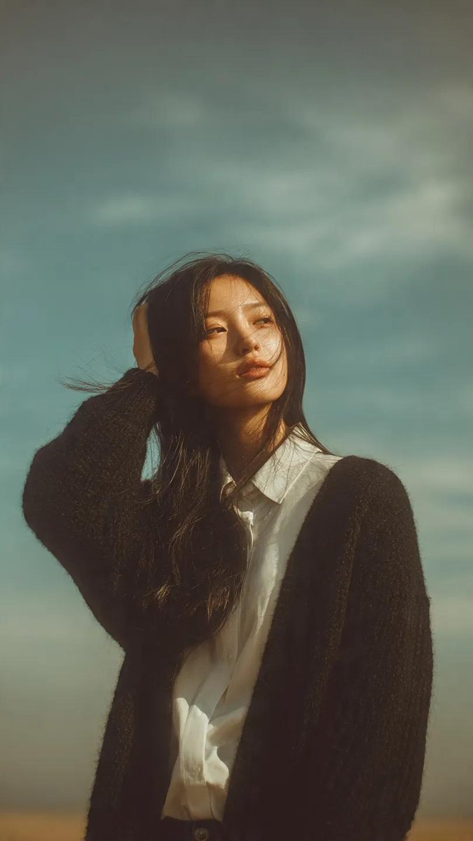一幅梦幻般的肖像画,一位年轻女子站在户外,头顶是柔和、广袤的蓝天。她那又长又直的黑发在风中轻轻飘扬,脸上带着柔和、怅惘的神情。她身穿一件黑色针织开衫,内搭一件挺括的白衬衫,显得既随性又不失时尚...