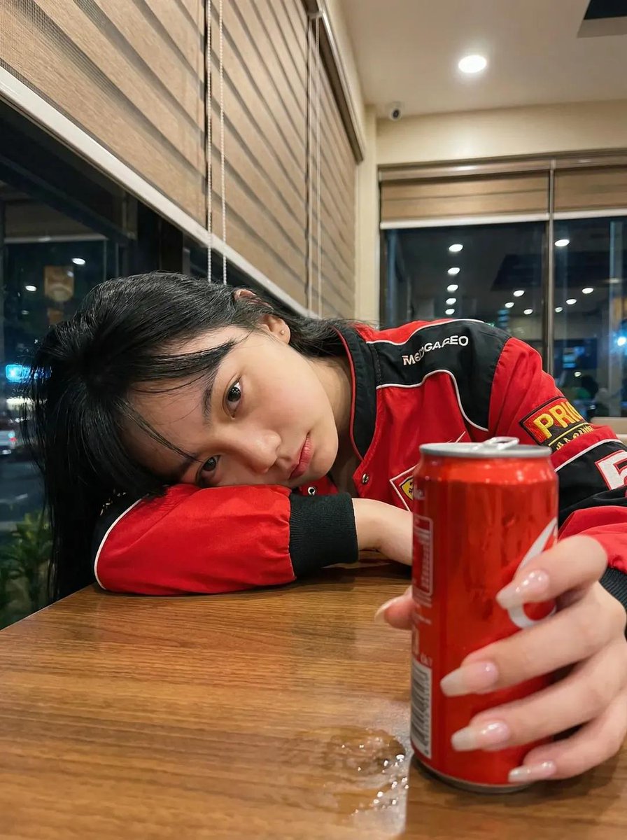 一张超写实的照片：一位年轻女性深夜坐在一间温馨的咖啡馆里，侧着头靠在自己放在木质桌面上的手臂上。她穿着一件红黑配色的赛车风格夹克。表情平静、略带疲惫、若有所思，直视镜头。一只手在前景中握着一罐...
