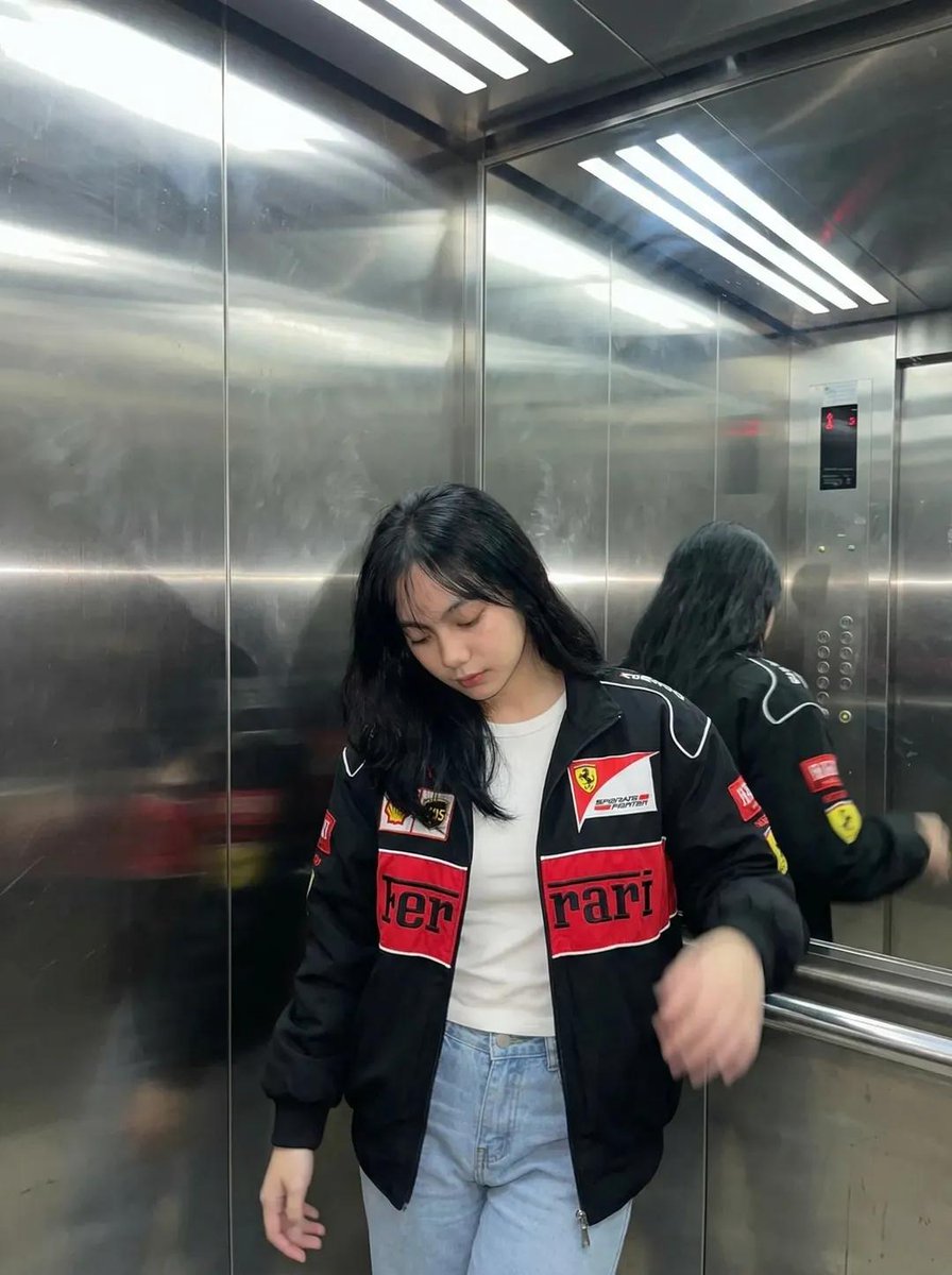一张超写实的照片：一位年轻女性深夜坐在一间温馨的咖啡馆里，侧着头靠在自己放在木质桌面上的手臂上。她穿着一件红黑配色的赛车风格夹克。表情平静、略带疲惫、若有所思，直视镜头。一只手在前景中握着一罐...
