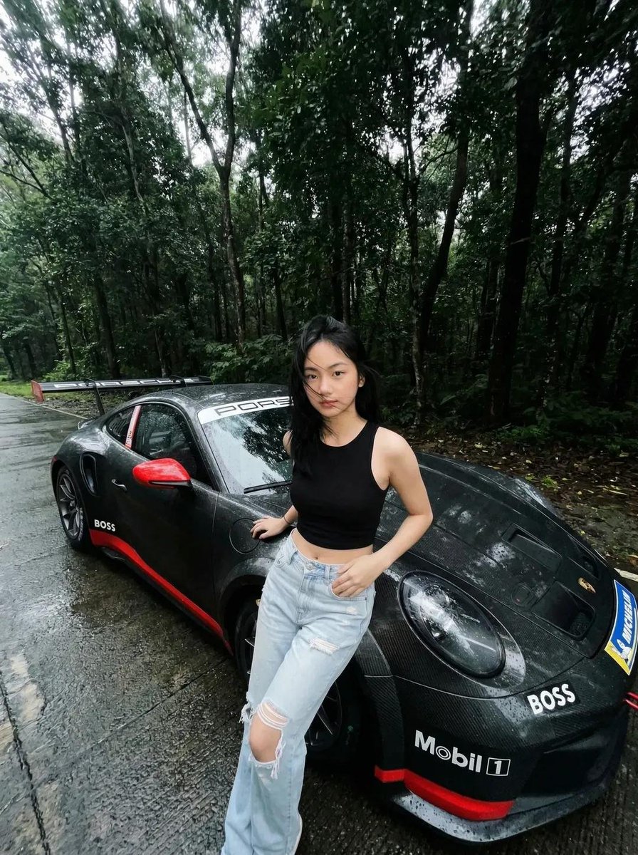 一张电影感、超写实的照片：一位年轻亚洲女性站在雨后湿润的森林道路上，旁边是一辆黑色保时捷跑车。她身穿黑色无袖短款上衣和浅蓝色破洞牛仔裤。她的头发略显凌乱，自然飘动，表情平静自信，直视镜头。 汽...