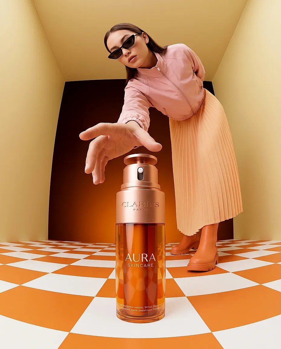 “超现实时尚产品摄影：一只巨大的琥珀色护肤品瓶（Clarins Aura）放置在一个极简盒子房间内的橙白格子地板上，温暖的纯橙色灯光，一位时尚女性戴着太阳镜、穿着桃色夹克伸手去够那只瓶子，广角...