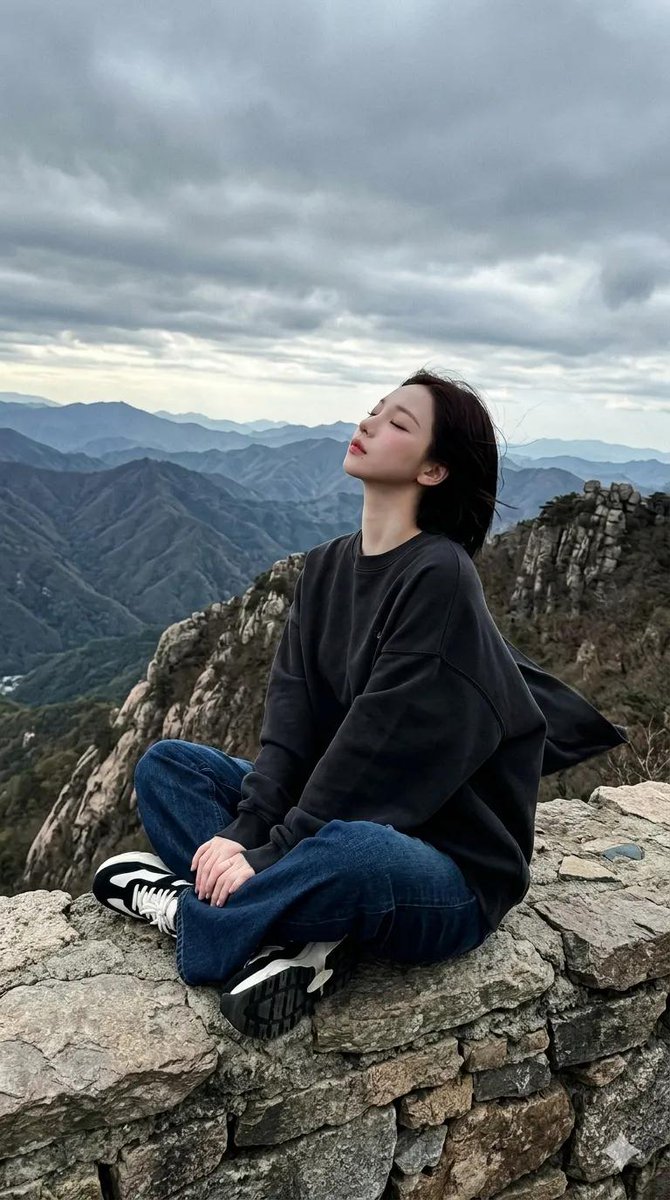 一段电影般的全景画面在戏剧性阴云笼罩的天空下，于一处嶙峋的山脊上徐徐展开。一位女性盘腿坐于石质岩架之上，身着超大号白色印花卫衣、深色阔腿牛仔裤以及黑白配色的厚底运动鞋。她头部微微后仰，双眼轻闭...