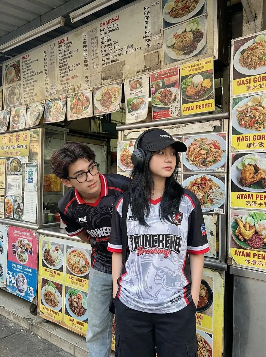 一张抓拍风格的街拍照片：两位年轻的亚洲成年人站在一家小型东南亚街边小吃摊前。小吃摊上挂满了色彩鲜艳的菜单板和食物照片，主打菜品包括 nasi goreng（印尼炒饭）和 ayam penyet...