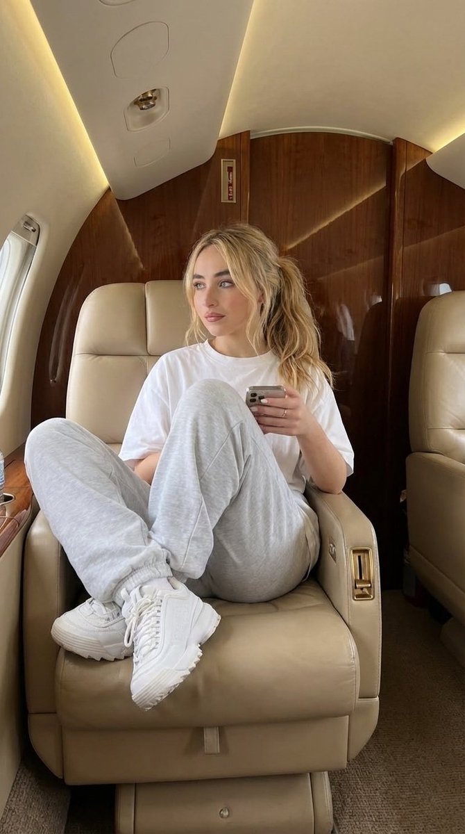 一位时尚年轻女性坐在私人飞机机舱内的全身照片,豪华内饰配有米色皮革座椅和抛光的木质面板,柔和温暖的环境光,服装:超大号白色T恤,宽松版型运动裤,厚底白色运动鞋,极简珠宝配饰,手持智能手机,姿势...
