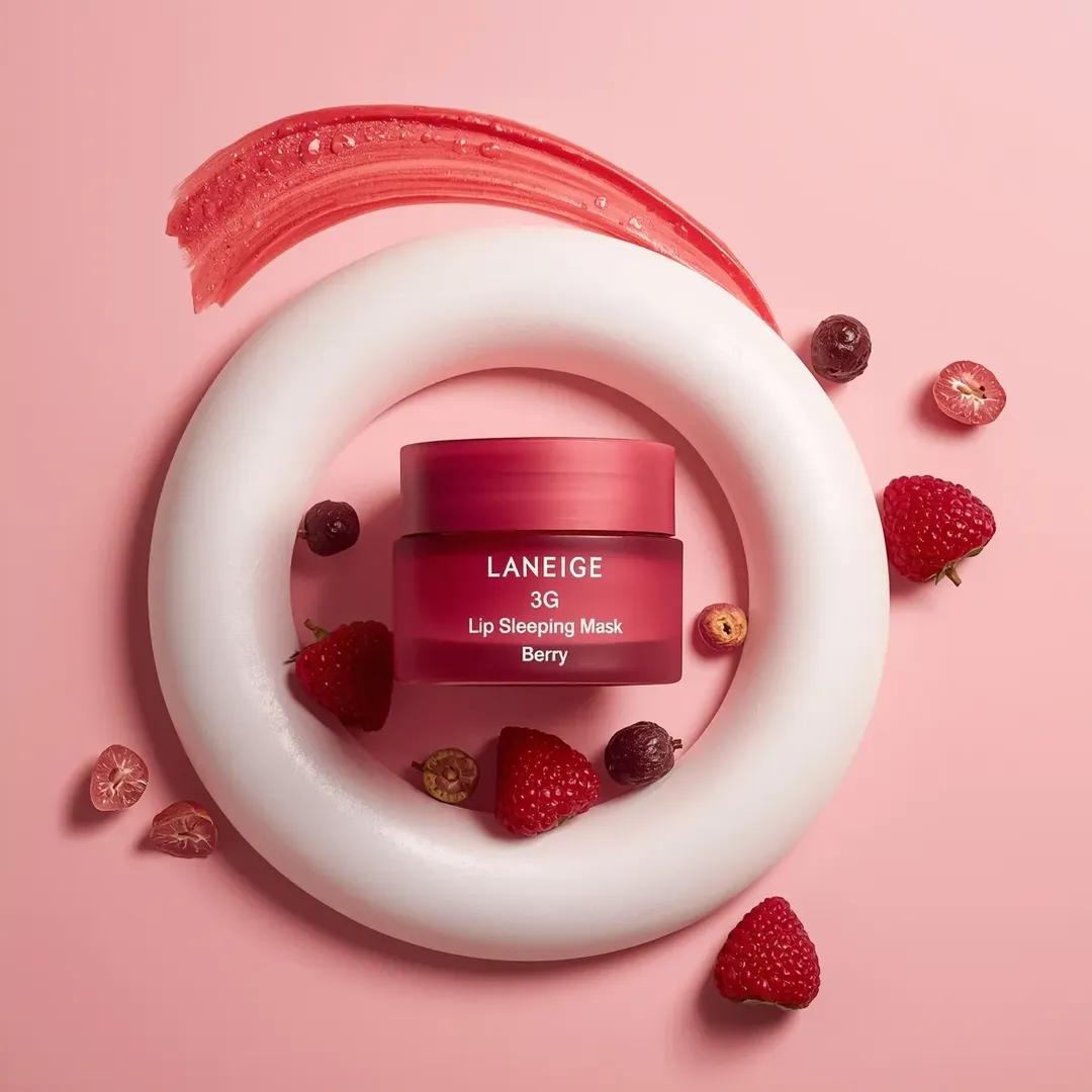一个粉色化妆品罐居于画面中央,标签为“LANEIGE Lip Sleeping Mask [Berry] 3 g”,放置在一大团旋涡状白色奶油之上。该罐子采用哑光半透明的粉色容器,配有极简风格...