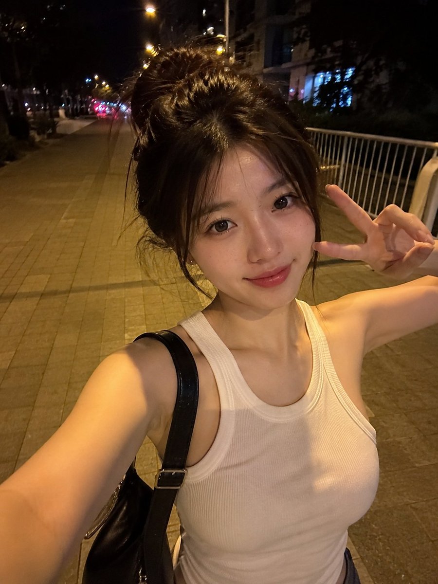 夜晚，一位美丽的东亚年轻女性在安静的城市人行道上户外行走的真实感智能手机自拍。她拥有瓷白肤色、闪亮的大眼睛呈深棕色、小巧笔挺的鼻梁、自然有光泽的粉嫩双唇，以及精致年轻的面部特征。她的深棕色头发...