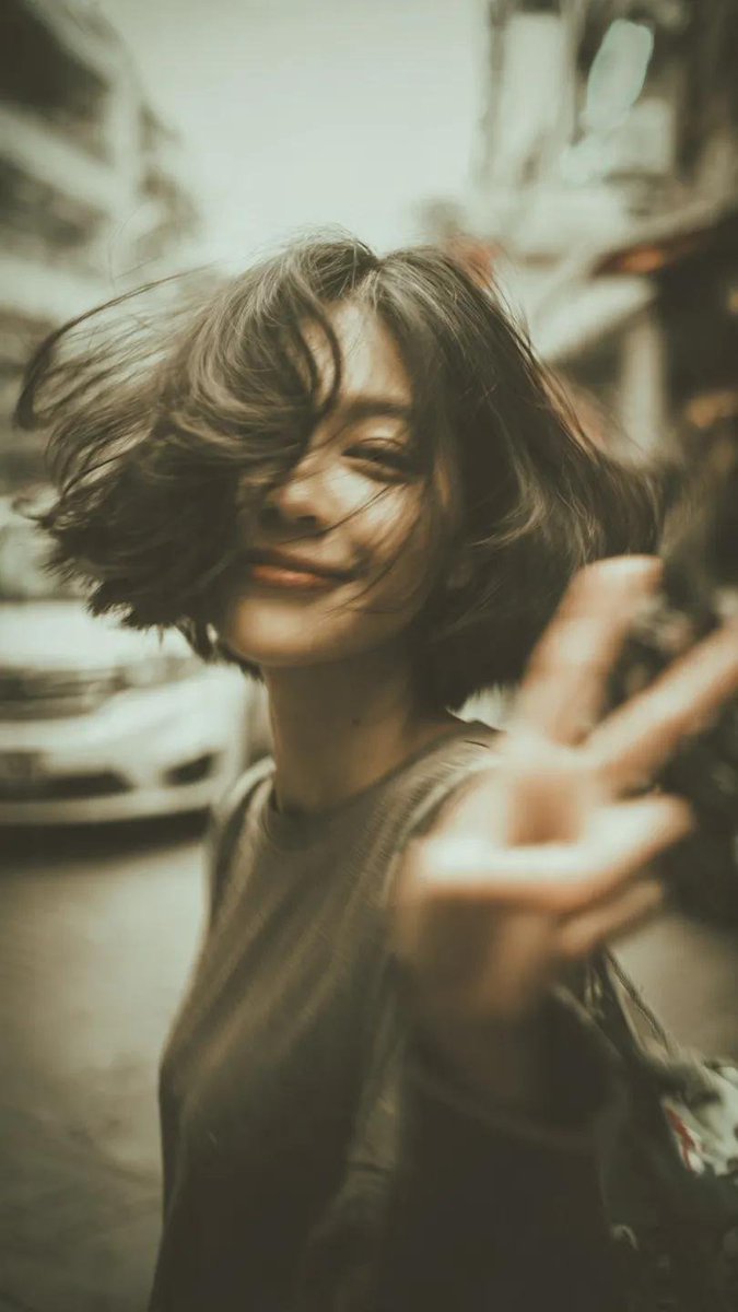 一张千禧年(Y2K)风格的街拍人像写真:一位开朗的年轻女性,留着凌乱的短波波头(bob),自然地开怀大笑,直接对着镜头比出胜利手势(V字手势/peace sign),她伸出的手占据前景,带有夸...