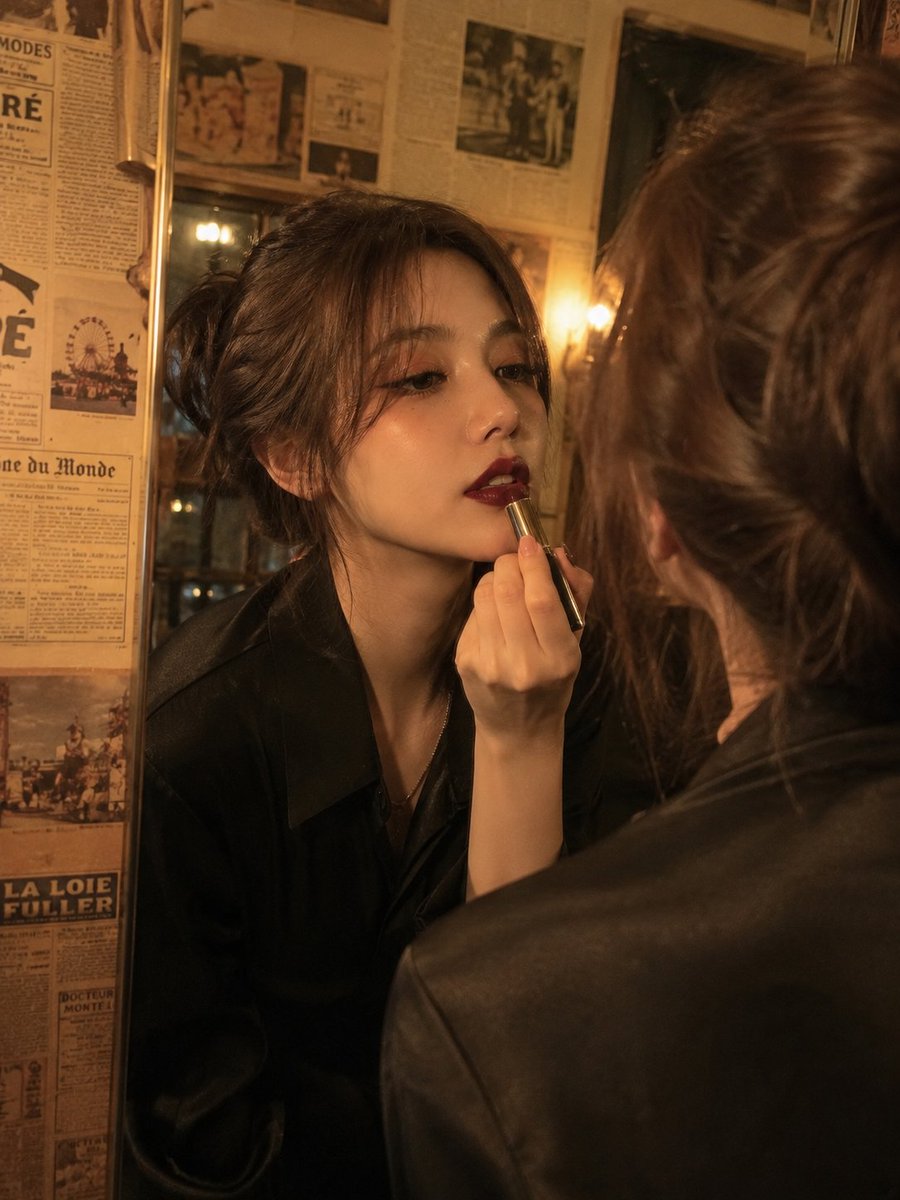 电影级复古美人肖像：一位优雅的年轻女性正在涂抹深酒红色口红，同时望向镜中，温暖的金色钨丝灯光在她脸上投下戏剧性的阴影，柔软的波浪棕色头发盘成略显凌乱的发髻，肌肤无瑕，妆容大胆，配以猫眼眼线和暗...