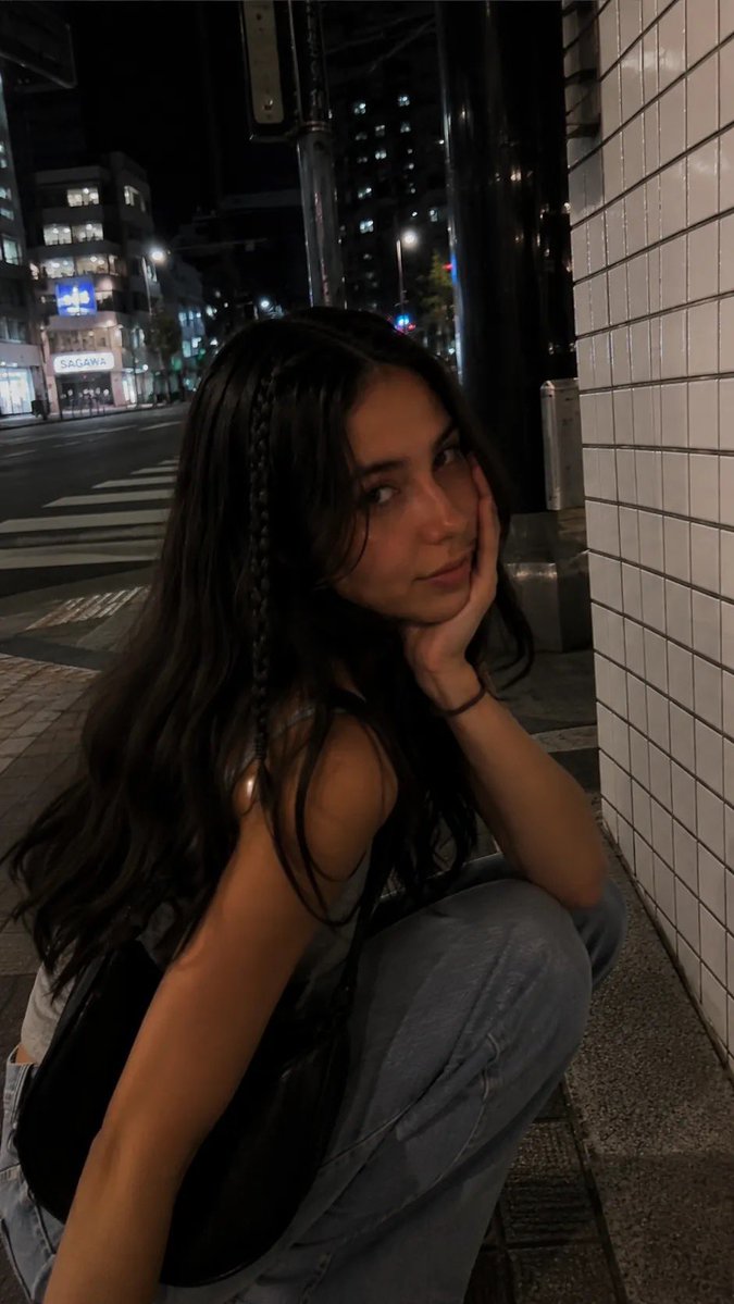 一段电影感十足的夜间街拍人像，写实抓拍风格，在日本城市安静的人行道上用 iPhone 拍摄。一位年轻女性沿着人行道行走，长而微卷的深棕色头发，一侧脸旁垂着一根细辫子，妆容自然柔和，微微带笑回头...