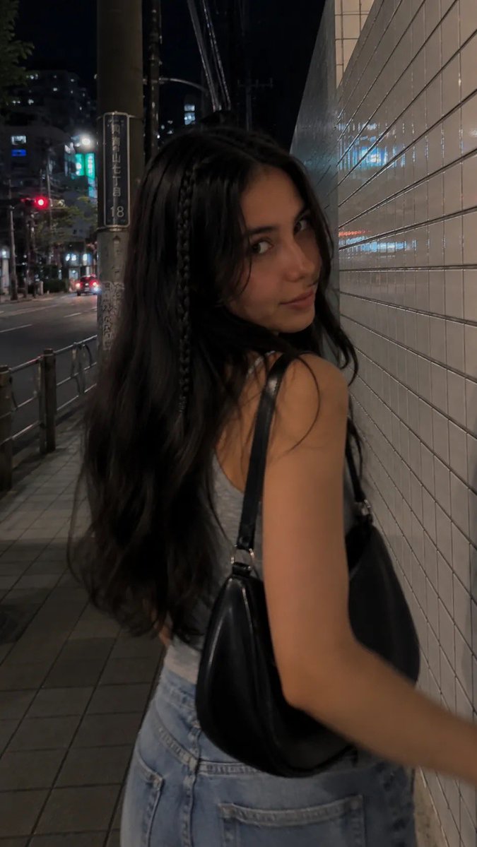 一段电影感十足的夜间街拍人像，写实抓拍风格，在日本城市安静的人行道上用 iPhone 拍摄。一位年轻女性沿着人行道行走，长而微卷的深棕色头发，一侧脸旁垂着一根细辫子，妆容自然柔和，微微带笑回头...