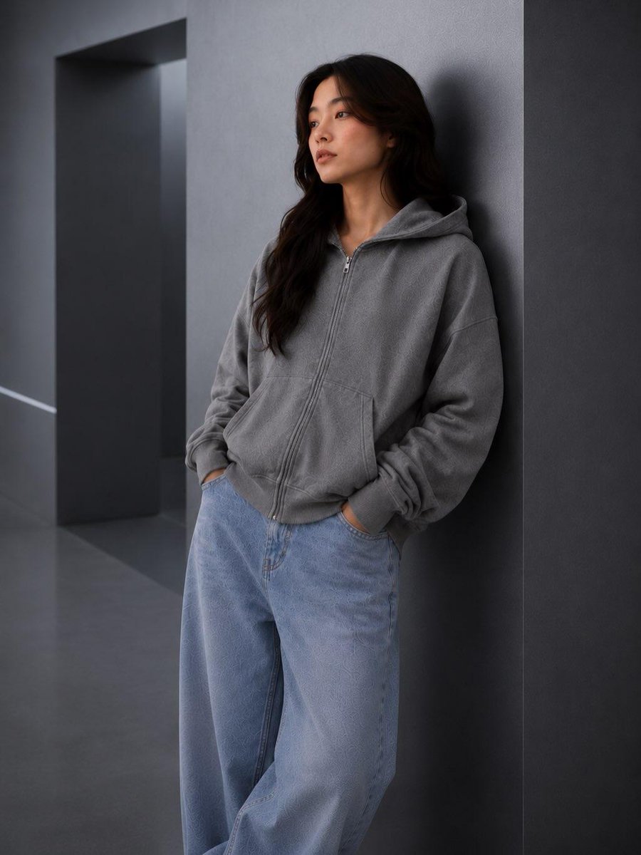 极简时尚编辑人像：一位年轻女性随意倚靠在一面光滑的炭灰色建筑墙面上，身穿水洗灰色宽松拉链连帽衫、浅色水洗宽松牛仔裤和白色运动鞋，双手插兜，姿态放松，神情平静自信，深色自然长卷发，淡妆自然，现代...