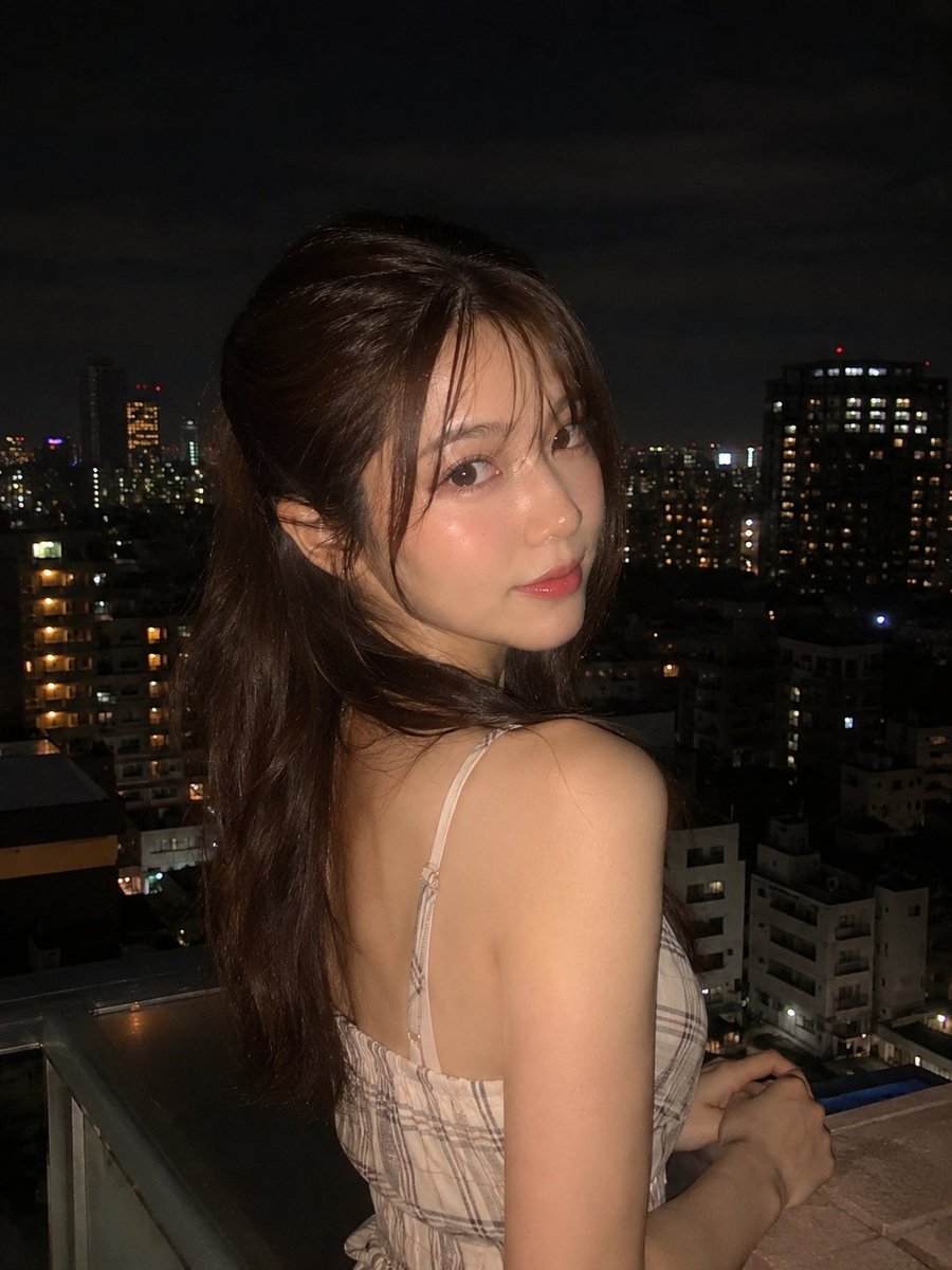 超写实夜景人像：一位美丽的年轻女子站在阳台上，俯瞰城市天际线，身穿优雅的无袖米色格纹连衣裙，淡雅腮红妆容，自然水润唇妆，慵懒的半扎发造型，几缕碎发自然垂落于脸颊两侧。她回眸望向镜头，神态温柔，...