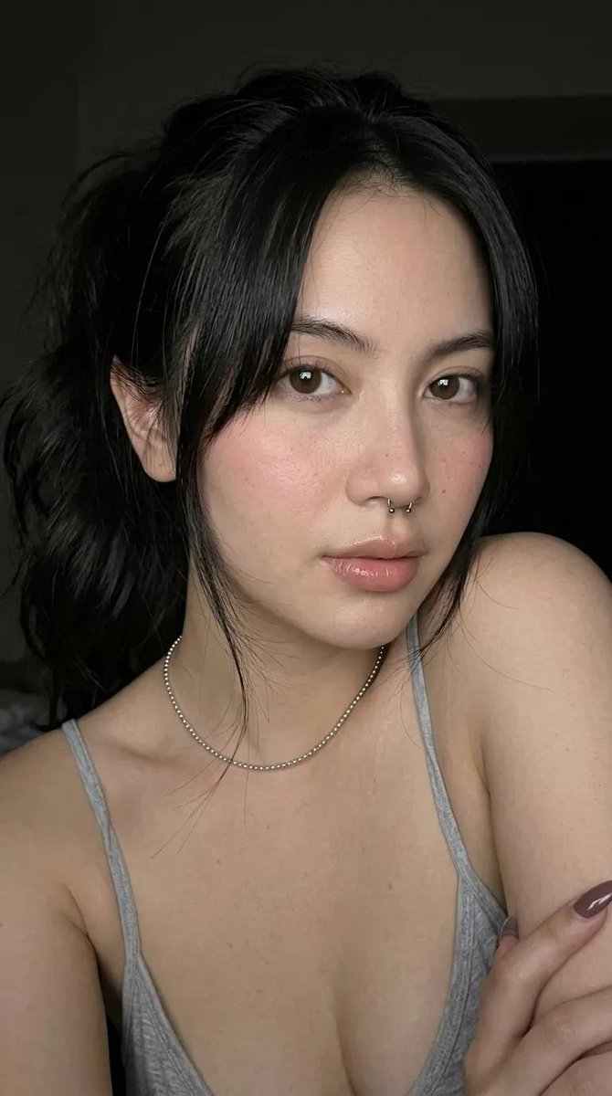 一张高端时尚工作室人像照，主体是一位年轻女性，肤色白皙，深棕色头发梳成光滑的高发髻。她呈紧凑的蹲姿，膝盖紧贴胸口。她平静、自信、神情平和地直视镜头。她身穿一件奶油色抹胸（心形领口）上衣，搭配宽...