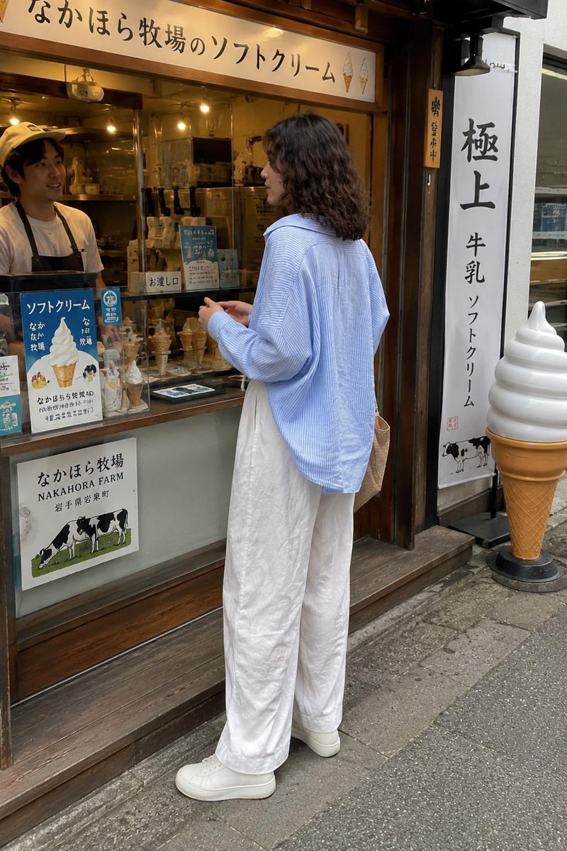 一张抓拍的旅行照片：一位年轻女子站在一家传统日式软冰淇淋店外，回眸对镜头微笑。她留着齐肩深色波浪卷发，身穿宽松的浅蓝色条纹纽扣衬衫、宽松的白色阔腿亚麻长裤和白色极简风格运动鞋，手里拎着一个米色...