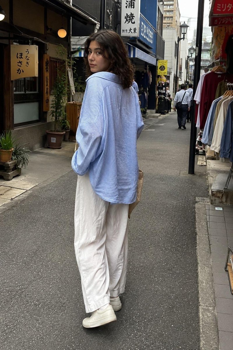 一张抓拍的旅行照片：一位年轻女子站在一家传统日式软冰淇淋店外，回眸对镜头微笑。她留着齐肩深色波浪卷发，身穿宽松的浅蓝色条纹纽扣衬衫、宽松的白色阔腿亚麻长裤和白色极简风格运动鞋，手里拎着一个米色...