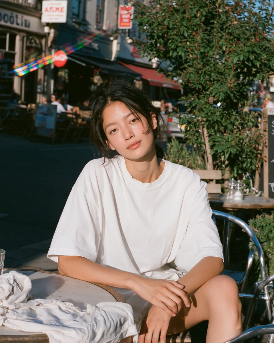 温暖阳光下的胶片人像，年轻女性放松地坐在小咖啡馆露台桌旁沐浴在明亮的金色午后阳光下，目光平静柔和、双眼睁开，身穿超大号白色T恤，身后是清晰可辨的户外街景，场景明亮通透，一束棱镜般的彩虹镜头光晕...