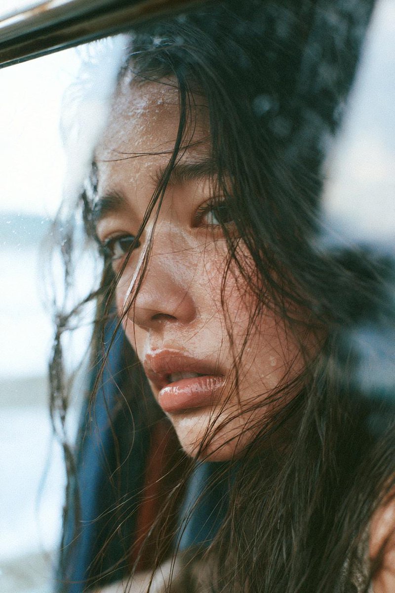 一位忧郁的年轻东亚女性，留着略显凌乱的长黑发，在黄金时刻静静坐在一扇大窗旁，柔和的阳光以温暖的电影色调照亮她的面庞。她若有所思地望向远方，妆容自然，皮肤水润，眼神富有表现力，几缕松散的头发在微...