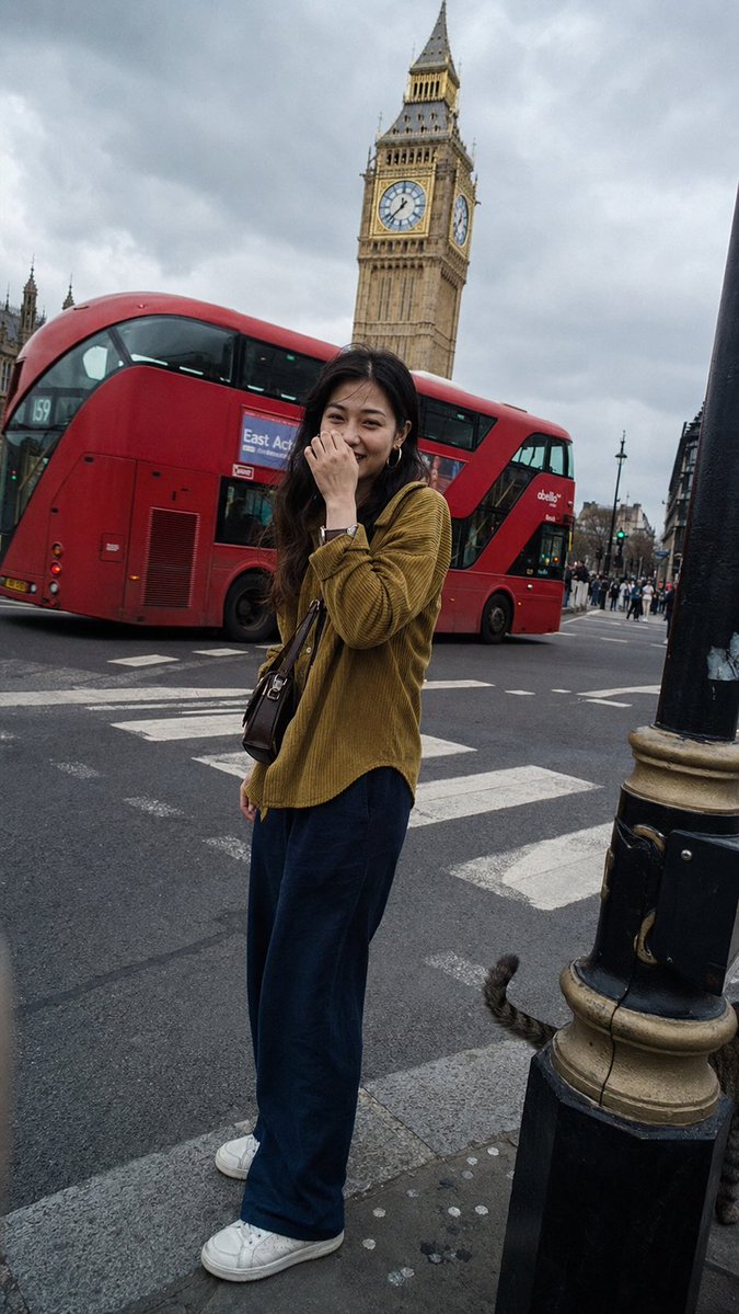 一张在伦敦街头随手拍摄的普通抓拍快照，看起来像是有人走在她前面，转身拍了一张照片。一位二十出头的年轻东亚女性站在人行横道前，她的容貌百分之百写实——直视镜头，带着疲惫却温暖真诚的笑容，眼睛因笑...