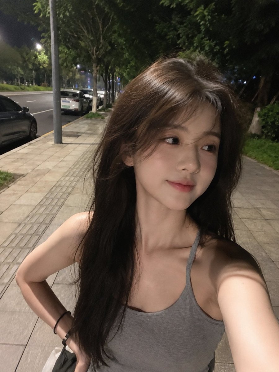 夜晚用手机拍摄的极致真实感智能手机自拍，看起来确实是用 iPhone/手机摄像头抓拍的，而不是单反相机。一位美丽的年轻东亚女性，留着浓密略带凌乱的深棕色长发，在微风中自然飘扬，拥有瓷白无瑕的肌...