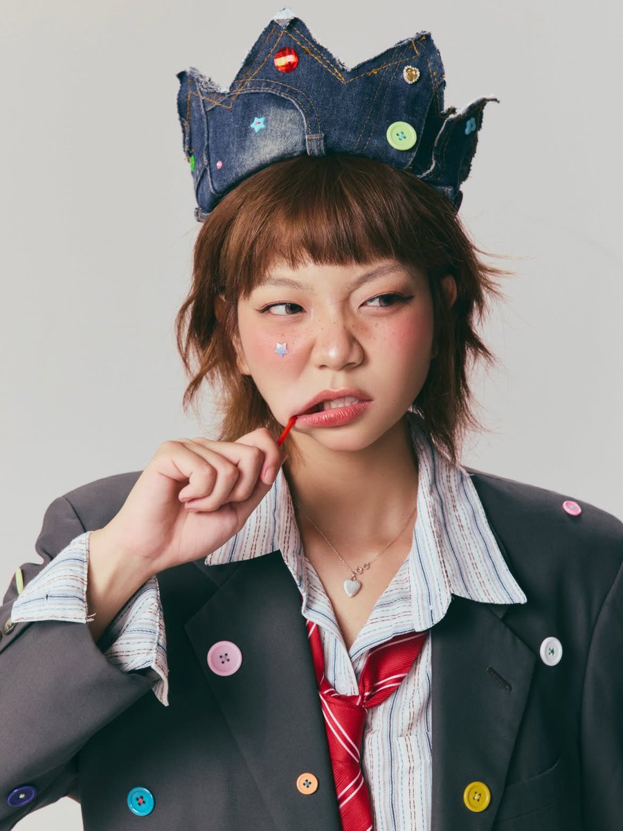 超写实编辑时尚肖像：一位俏皮的年轻女性，留着短款层叠栗棕色波波头与齐整的直刘海，柔和自然妆容配以玫瑰色腮红，鼻梁上有淡淡的雀斑，粉色光泽唇，长而卷翘的睫毛，搭配上挑眼线，一侧面颊贴着一枚小巧的...