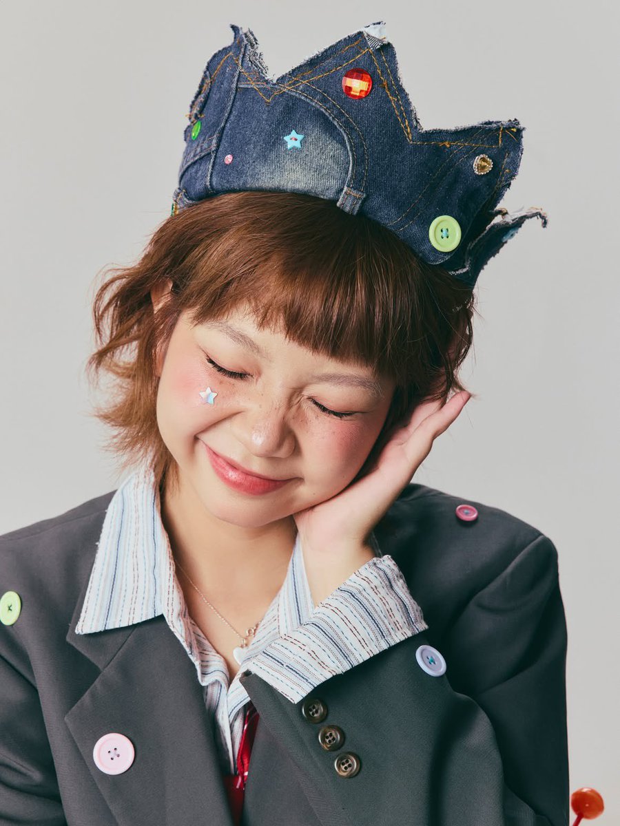超写实编辑时尚肖像：一位俏皮的年轻女性，留着短款层叠栗棕色波波头与齐整的直刘海，柔和自然妆容配以玫瑰色腮红，鼻梁上有淡淡的雀斑，粉色光泽唇，长而卷翘的睫毛，搭配上挑眼线，一侧面颊贴着一枚小巧的...