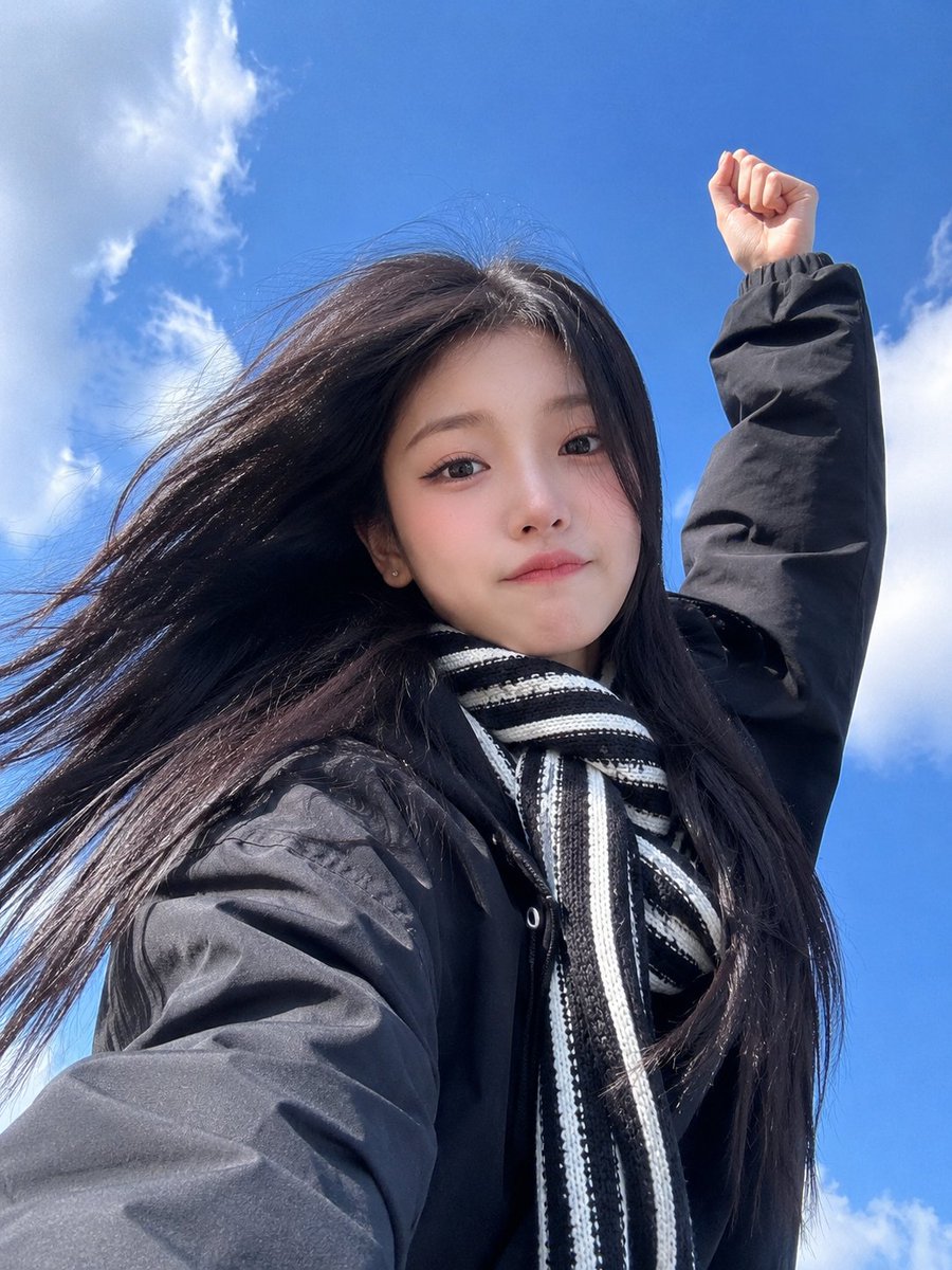 极致真实的智能手机自拍照，一位美丽的东亚年轻女性，从极具戏剧性的低角度仰拍，背景是鲜艳的蓝天和柔和的白色云朵。她拥有又长又直的乌黑长发在风中自然飘动，白皙的瓷器肌肤，柔和的杏仁形棕色眼睛，细腻...