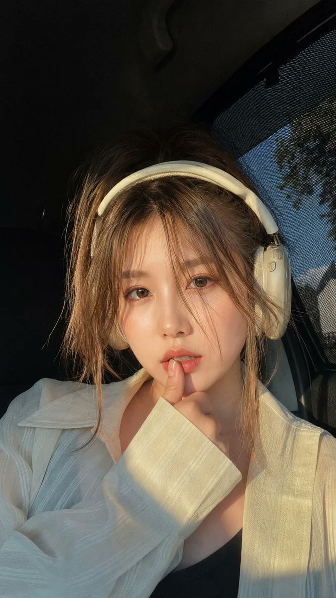 超写实特写肖像：一位美丽的年轻女性坐在车内,正值黄金时刻,戴着优雅的米白色头戴式耳机。柔和的自然阳光从侧窗洒入,在她的脸庞和宽松的白色衬衫上投射出温暖的电影感高光。她拥有白皙瓷肌、温暖的大棕色...