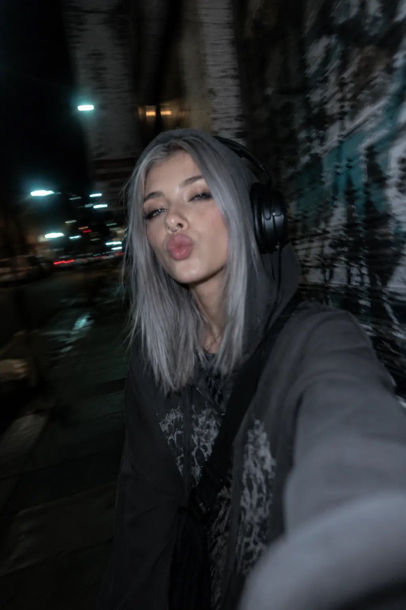 以所附照片为参考，创作一张夜间城市摄影人像，主体为一位年轻女性，整体呈现另类、垃圾摇滚（grunge）美学。她留着齐肩直发、中分，浅色眼睛搭配浓重的黑色猫眼眼线、纤长睫毛，戴着鼻钉，嘴唇微启且...