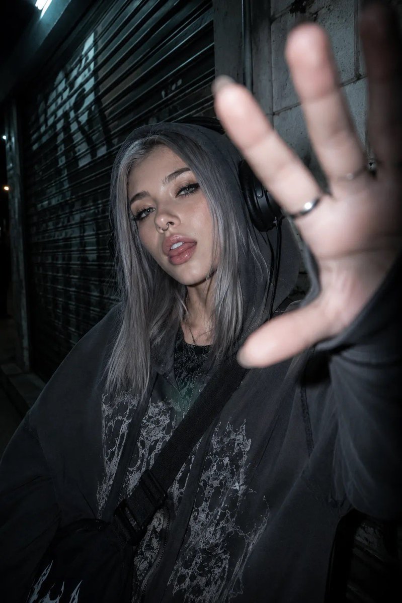 以所附照片为参考，创作一张夜间城市摄影人像，主体为一位年轻女性，整体呈现另类、垃圾摇滚（grunge）美学。她留着齐肩直发、中分，浅色眼睛搭配浓重的黑色猫眼眼线、纤长睫毛，戴着鼻钉，嘴唇微启且...