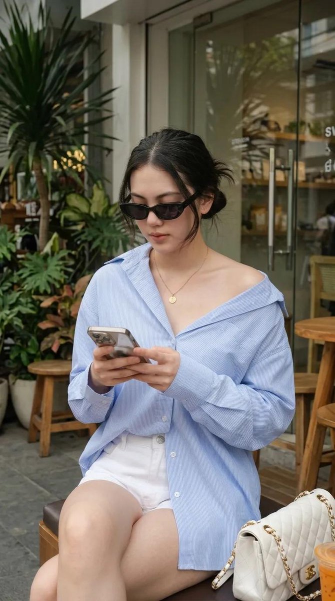 使用我的图片，生成一张 9:16 的图像 --- 一位年轻女性坐在户外咖啡馆，随性慵懒的 Oversized 风格。身穿宽松 Oversized 浅蓝色细条纹纽扣衬衫，袖子卷起，衬衫垂落足够长...