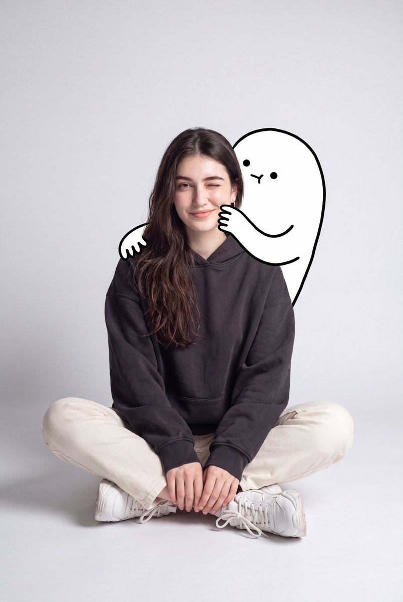 一幅可爱的极简风肖像：一位年轻女性随意地坐在地板上，身处一个光线柔和的灰色背景的简洁摄影棚中。她穿着一件宽松的深炭灰色连帽衫、奶白色直筒长裤、白色袜子和厚底白色运动鞋。她盘腿而坐，姿态放松，一...