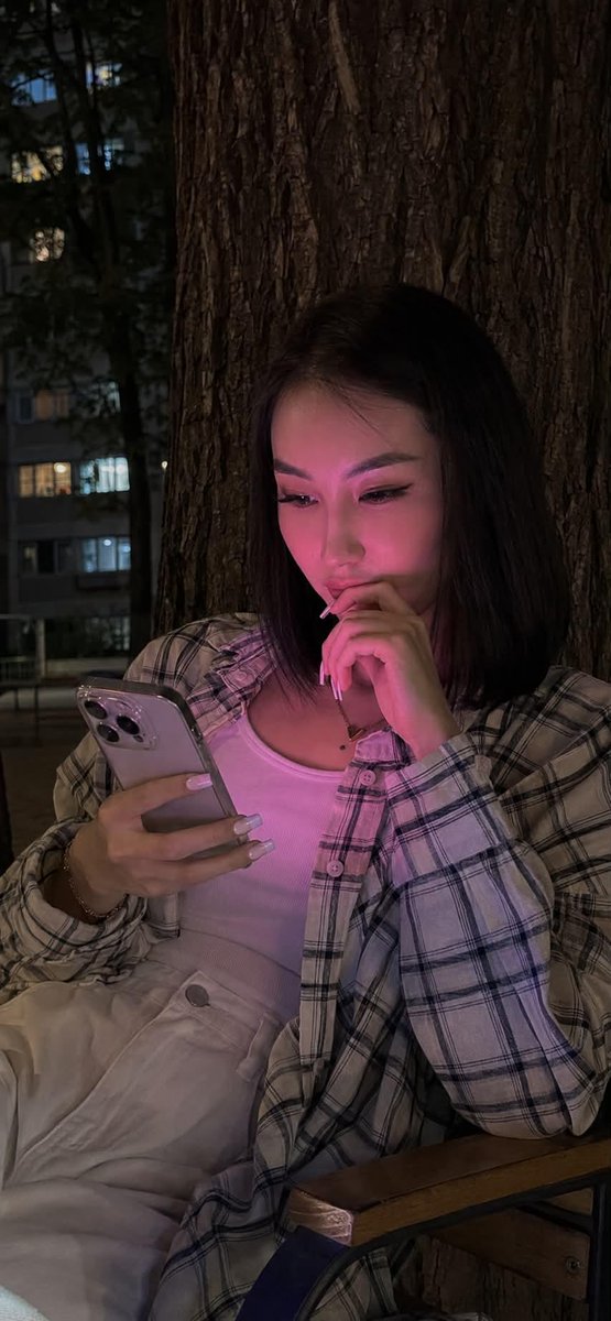 夜晚使用 iPhone 15 Pro 拍摄的写实风格生活照。画面中央是一位年轻女性，留着及腰的黑色长直发。她正饶有兴趣地注视着智能手机，嘴角轻轻上扬，温柔地微笑。 妆容：长簇式睫毛嫁接，眉形清...