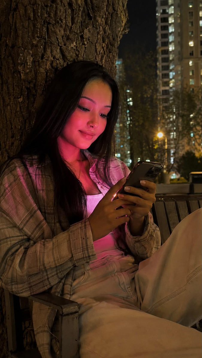 夜晚使用 iPhone 15 Pro 拍摄的写实风格生活照。画面中央是一位年轻女性，留着及腰的黑色长直发。她正饶有兴趣地注视着智能手机，嘴角轻轻上扬，温柔地微笑。 妆容：长簇式睫毛嫁接，眉形清...