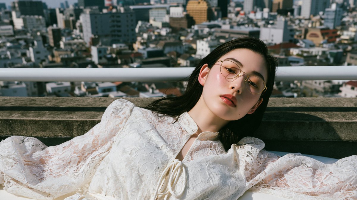 一位惊艳的二十多岁日本或韩国女性，戴着金色金属细框眼镜，身穿白色蕾丝泡泡袖系带领口衬衫，以自然躺卧的姿势斜靠在床沿；头部转向镜头，位于高楼屋顶边缘，背景有屋顶护栏和混凝土女儿墙，在正午强烈光线...
