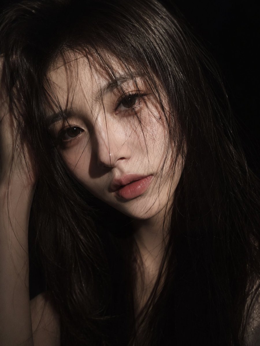 一位东亚年轻女性的电影感特写肖像，长而略显凌乱的黑发散落在脸庞，瓷器般白皙的肌肤，柔和的粉色双唇，以及深邃而富有表现力的双眸。她以平静、忧郁的神情直视镜头。戏剧化的低调照明仅照亮她面部的部分区...