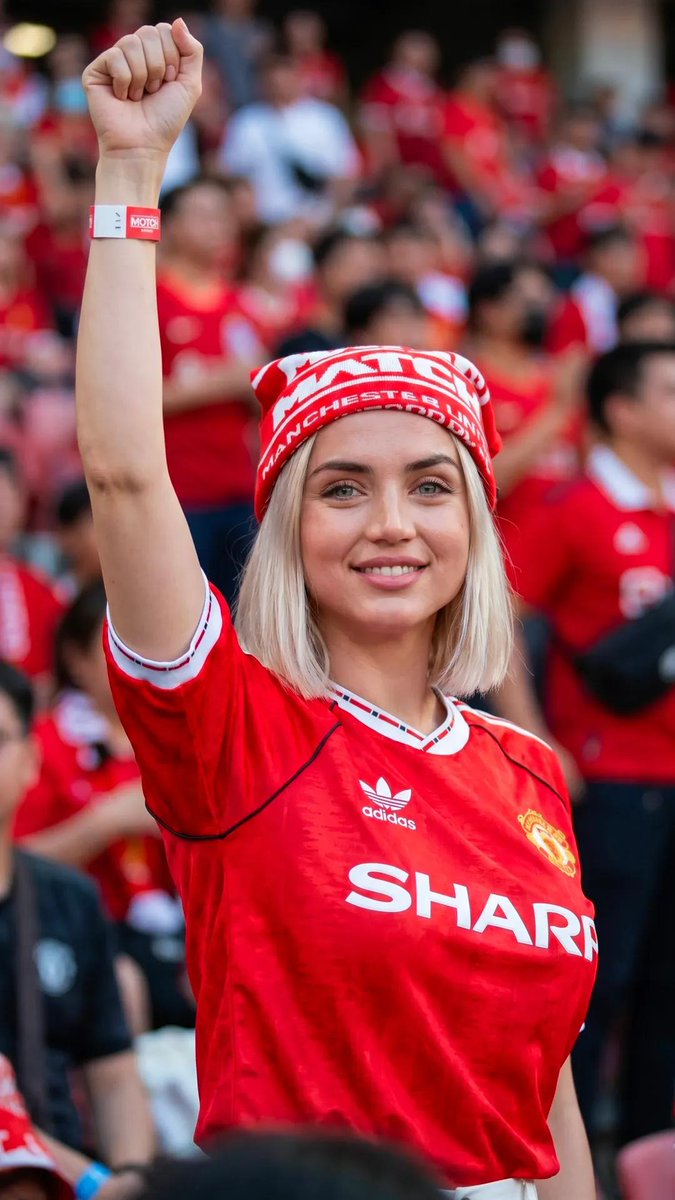 超现实的体育生活方式编辑风格人像：一位惊艳的女性足球迷，留着利落的铂金金色波波头发型，眼神呈冰蓝偏绿色，皮肤纹理自然，妆容柔和，神态自信。她身穿复古款红色曼联球衣（1990年代 SHARP 赞...