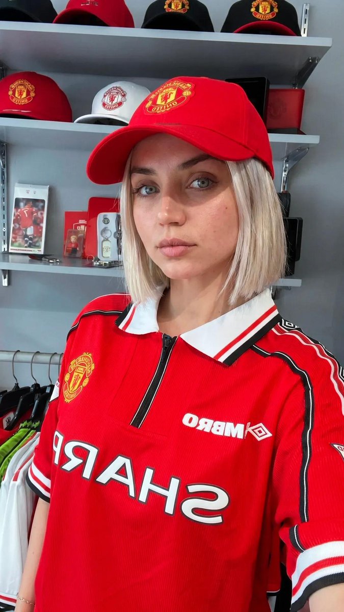 超现实的体育生活方式编辑风格人像：一位惊艳的女性足球迷，留着利落的铂金金色波波头发型，眼神呈冰蓝偏绿色，皮肤纹理自然，妆容柔和，神态自信。她身穿复古款红色曼联球衣（1990年代 SHARP 赞...