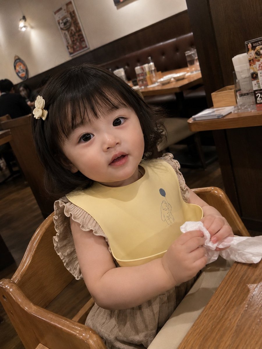 真实感抓拍智能手机照片：一位可爱的女幼儿坐在一家温馨的家庭餐厅的木制高脚椅上。她有着柔软的刘海短发，戴着一个小小的奶白色发夹，眼睛又大又有神，脸颊红润白皙。她身穿浅棕色荷叶边上衣，围着一条柔和...