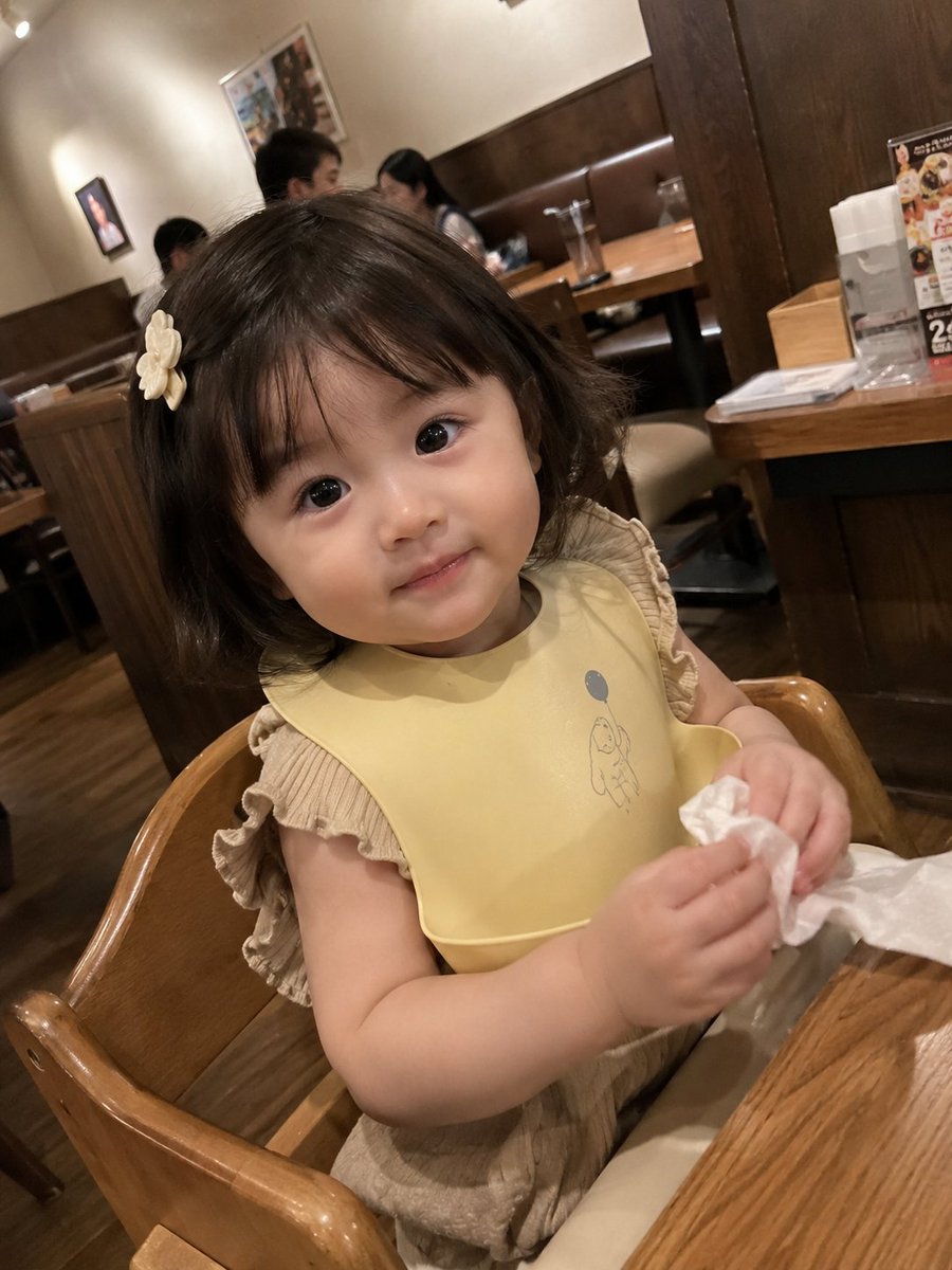 真实感抓拍智能手机照片：一位可爱的女幼儿坐在一家温馨的家庭餐厅的木制高脚椅上。她有着柔软的刘海短发，戴着一个小小的奶白色发夹，眼睛又大又有神，脸颊红润白皙。她身穿浅棕色荷叶边上衣，围着一条柔和...