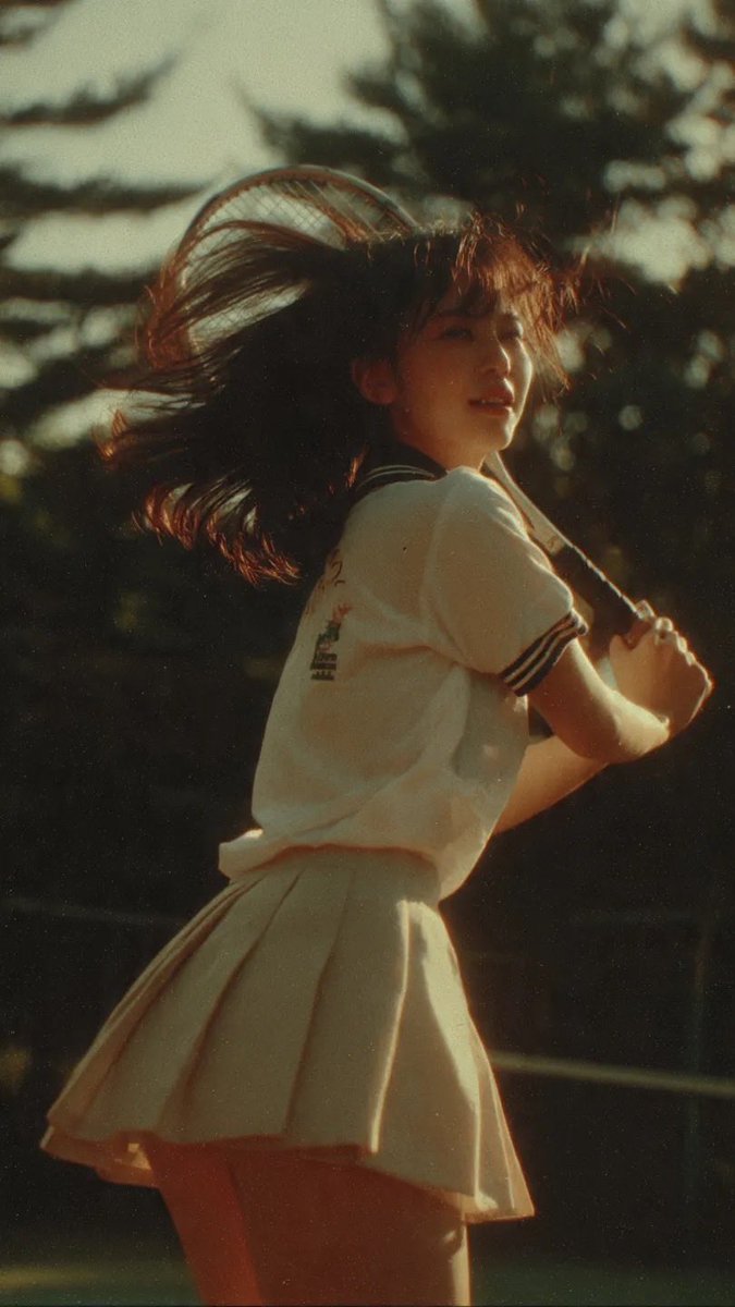 电影感中景镜头：一名身穿网球服的日本少女，在网球比赛挥拍途中被自然抓拍，柔和阳光穿过树林，暖色调，90年代日本电影美学，轻微胶片颗粒感，风吹动她的头发，动态而自然的动作，怀旧诗意氛围。