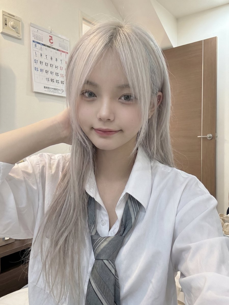 一位年轻的日本女孩，留着长长的银白色头发，柔软凌乱的刘海部分遮住眼睛，侧面编着一条用粉色丝带扎住的小辫子，穿着白色纽扣校服衬衫、灰色领带和校服。她随意地坐在室内，一只手托着脸，用平静、略带困倦...
