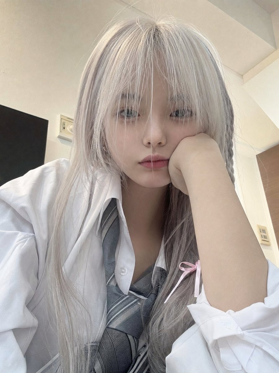 一位年轻的日本女孩，留着长长的银白色头发，柔软凌乱的刘海部分遮住眼睛，侧面编着一条用粉色丝带扎住的小辫子，穿着白色纽扣校服衬衫、灰色领带和校服。她随意地坐在室内，一只手托着脸，用平静、略带困倦...