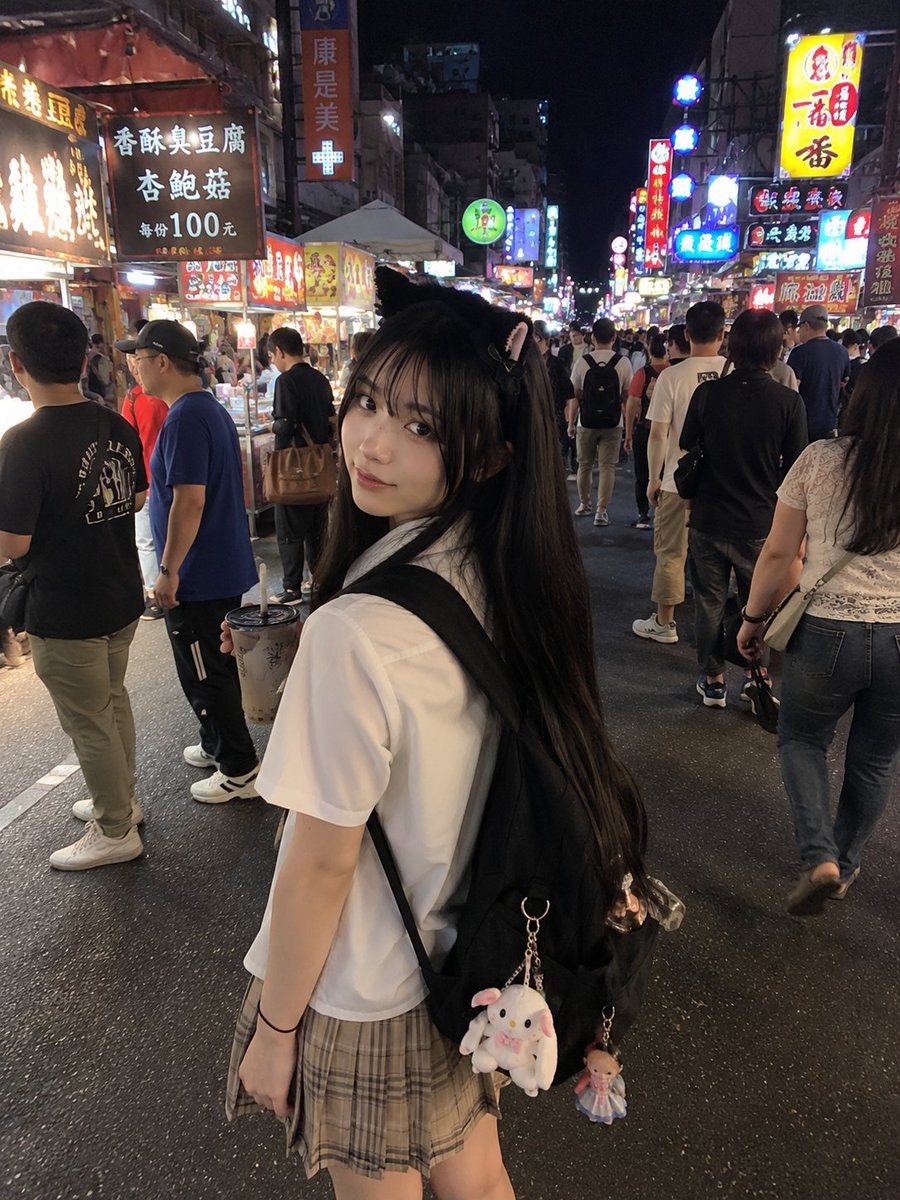 年轻的日本女孩，留着顺直黑色长发和柔软的刘海，戴着可爱的黑色猫耳发箍，身穿白色短袖衬衫、条纹领带和米色格纹百褶裙。她站在一个充满活力的亚洲夜市里，四周是霓虹招牌、美食摊位和熙熙攘攘的人群。她一...