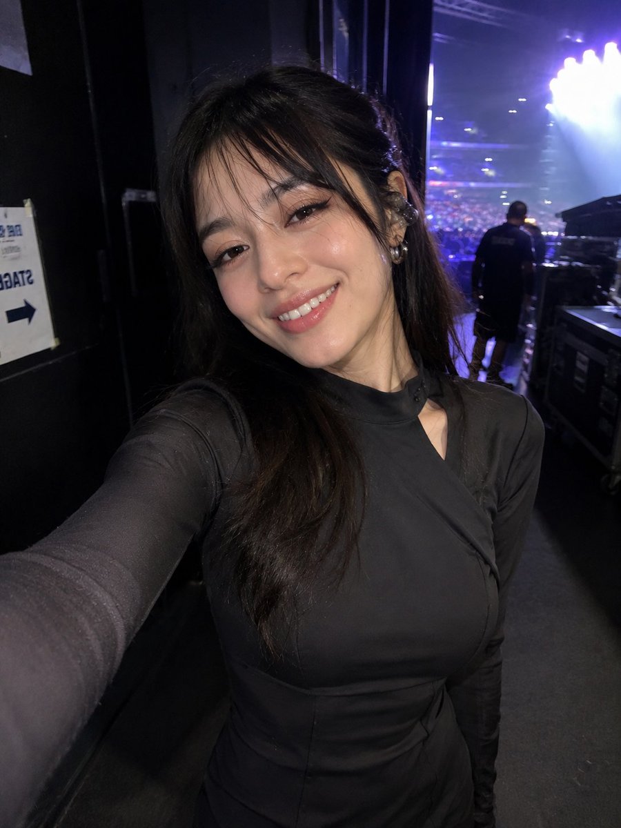 参考图片中同一女性的超写实偶然iPhone前置摄像头自拍，保留完全一致的身份、面部特征、发型和比例。在一场现场演唱会的后台，在她走过走廊时以抓拍方式捕捉。她微微转身，带着放松、演出后疲惫的微笑...