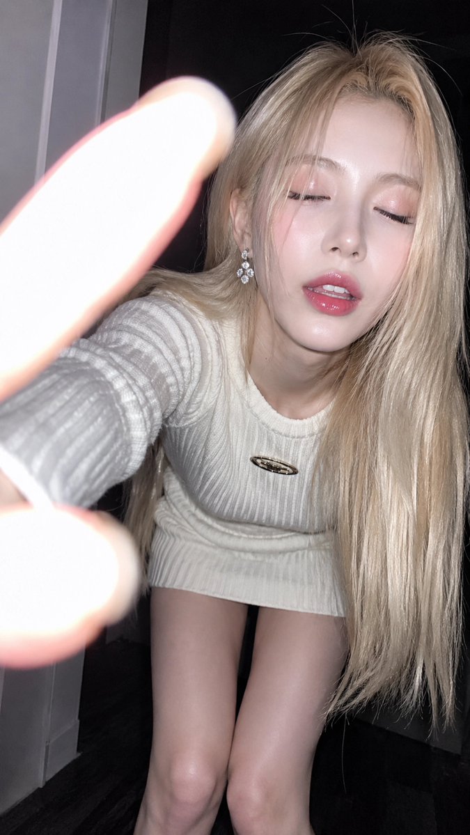 一名18岁以上的年轻成年女性，室内近距离闪光灯特写肖像。她向镜头前倾，营造出一种自然的自拍式瞬间。视觉形象由铂金色头发、白色罗纹针织迷你连衣裙、强烈闪光灯和深色室内背景构成, 不是镜子自拍；没...