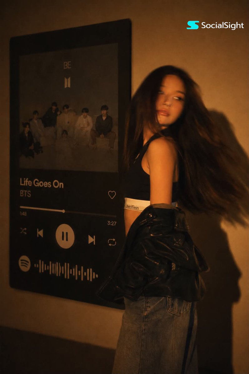 一位发型飘逸的深色长发时尚年轻女子在巨型壁挂Spotify音乐播放器显示屏前摆姿势。温暖的琥珀色电影级灯光布满房间，在墙上投下戏剧性阴影。她身穿黑色卡尔文·克莱因露脐上衣，廓形黑色皮夹克滑落肩...